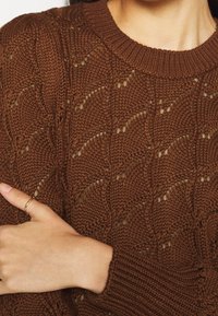 Brązowy sweter z dzianiny o strukturalnym wzorze, z okrągłym dekoltem i ściągaczami. Dzianina ma ażurowy wzór i miękką tkaninę.