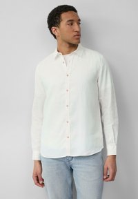 Chemise blanche en lin à manches longues avec col boutonné, présentant des boutons marron clair et un ourlet incurvé. Associée à un jean bleu.