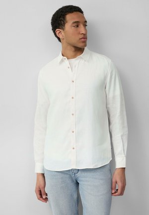 Chemise blanche en lin à manches longues avec col boutonné, présentant des boutons marron clair et un ourlet incurvé. Associée à un jean bleu.