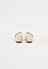 Pantofi din piele bej cu toc mic, spate deschis și un accent mic sub formă de fundiță pe lateral, având o textură netedă și un design minimalist.