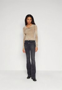 Smėlio spalvos ilgomis rankovėmis palaidinė su mazgo detalėmis ties kaklu, derinama su tamsiais, platėjančiais džinsais ir juodais aulinukais. Lygi audinio tekstūra.