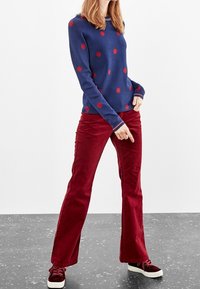 Maglione blu navy con pois rossi, abbinato a pantaloni a zampa di elefante in corduroy bordeaux. Modello che indossa sneakers rosso scuro con suole bianche.