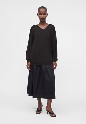 Femme debout portant un pull noir à col en V manches longues, une jupe midi noire et des sandales à talons ouvertes noires sur un fond uni.