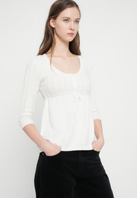 Λευκή πλεκτή μπλούζα με στρογγυλή λαιμόκοψη, κουμπιά στο μπροστινό μέρος και ζώνη με κορδόνι στη μέση. Μανίκια τριών τετάρτων με ριμπ τελειώματα.