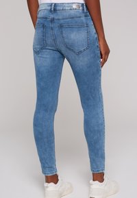 Jeans skinnys de denim azul con un acabado desgastado, que cuentan con bolsillos traseros y se combinan con zapatillas blancas, mostrando un diseño elegante.