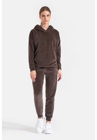 Brązowa bluza z kapturem z weluru z przednią kieszenią, zestawiona ze spodniami zwężanymi. Posiada miękką fakturę i elastyczne mankiety. Modelka nosi białe trampki.