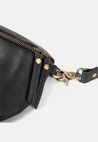 Schwarze Ledertasche mit doppelten Messingreißverschlüssen und passenden Zuglaschen, befestigt an einem Messing-Schwenkarabiner an einem schwarzen Lederband.