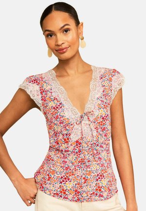 Nő, aki virágmintás blúzt visel csipkés díszítéssel a nyakkivágásnál és vállkendővel, krémszínű nadrággal párosítva.