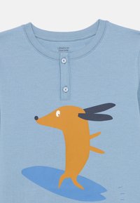 Μπλε πουκάμισο από οργανικό βαμβάκι με λαιμόκοψη henley, με ένα παιχνιδιάρικο πορτοκαλί σκύλο σε ανοιχτό μπλε φόντο.