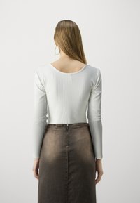 Top bianco a maniche lunghe con costine e schiena bassa, abbinato a una gonna di jeans marrone a vita alta con zip centrale e texture liscia.