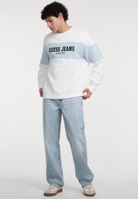 Felpa bianca con una striscia azzurro chiaro sul petto, con la scritta "GUESS JEANS" stampata in nero. Abbinata a jeans azzurri chiari e sneaker casual.