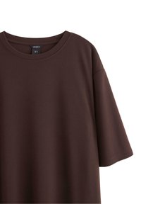 Brun T-shirt med korte ærmer, rund halsudskæring, glat tekstur og afslappet pasform. Mærket synligt nær kraven.