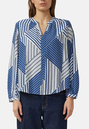 Kobieta ubrana w luźną, niebiesko-białą bluzkę z długim rękawem z geometrycznym paskowanym wzorem oraz ciemnoniebieskie jeansy, stojąca na tle białego tła.