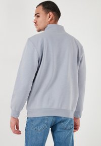 Sudadera de cuello alto en gris claro, con mangas largas, puños y dobladillo acanalados; combinada con jeans azules. Textura suave, ajuste casual.