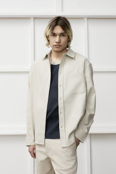 Chemise boutonnée beige clair avec une poche, portée sur un haut bleu foncé. Tissu uniforme, coupe décontractée, présentée sur fond blanc.