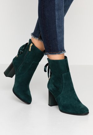 Buty za kostkę z zielonego zamszu w ciemnym odcieniu, na blokowym obcasie, z suwakiem z boku i kokardką z tyłu, noszone z ciemnoniebieskimi skinny jeansami z postrzępionymi brzegami.