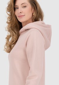 Hoodie rosa chiaro realizzato in un materiale morbido, con cappuccio regolabile e una texture liscia. Esposto su uno sfondo neutro.