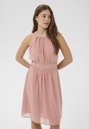 Młoda kobieta o długich brązowych włosach, ubrana w różową sukienkę bez rękawów z koronkowym pasem, stoi z rękami za plecami, patrząc na bok.