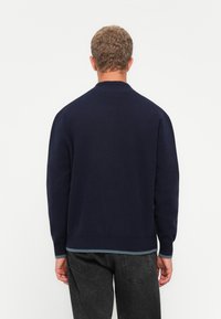 Man met kort krullend haar, gekleed in een marineblauwe trui en zwarte spijkerbroek, met zijn rug naar de camera gekeerd tegen een effen lichte achtergrond.