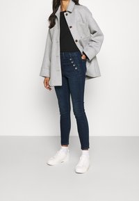 Ljusgrå jacka i ullblandning med bruna knappar, svart topp och mörkblå skinny jeans med sidoknappdetalj; vita sneakers fulländar looken.