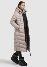 Long beige puffer coat with a hood, quilted design, front pockets, and black accents. Worn with black high-top sneakers.