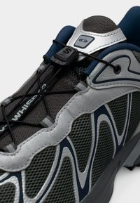 Ελαφρύ αθλητικό παπούτσι με γκρι δίχτυ επάνω μέρος, πράσινες λεπτομέρειες, ασημί επικαλύψεις και μαύρο σύστημα κορδονιών με κλείδωμα για σταθερή εφαρμογή.