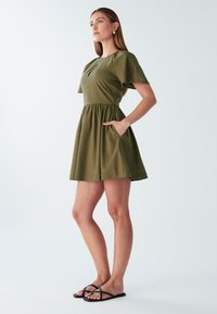 Olivno zeleni kratek rokav oblekica z oprijetim telovnikom in razširjenim krilom. Ima žepke in okroglo vratno izreza. Nosi se z črnimi sandalami.
