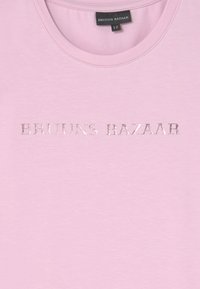 Camiseta de algodão de cor rosa claro com decote redondo e mangas curtas, apresentando texto em relevo prateado "BRUUNS BAZAAR" na parte da frente.