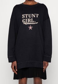 Svart oversized sweatshirt med grafik "STUNT GIRL" i beige, med en stjärnaccent och ribbade muddar. Bärs över en svart plisserad kjol.