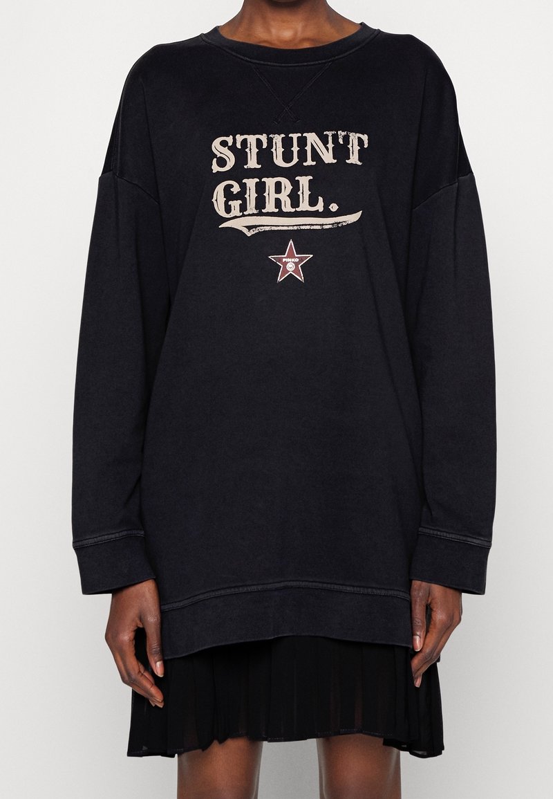 Svart oversized sweatshirt med grafik "STUNT GIRL" i beige, med en stjärnaccent och ribbade muddar. Bärs över en svart plisserad kjol.