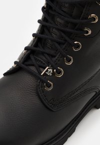 Botín de cuero negro con ojales de metal, cordones negros y costuras reforzadas. Suela de goma duradera y acabado texturizado.