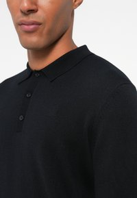 Črna pletena polo majica z ovratnikom, zapenjanjem na tri gumbe in subtilnim embosiranim logotipom na prsih. Gladka tekstura in prilega zasnova.