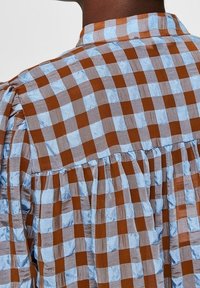 Ruskea ja vaaleansininen ruudullinen paita, jossa on kaulus, laskostettu selkä ja tekstuuri, joka antaa hieman kiiltoa.