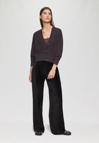 Dunkelgraue, verkürzte Strickjacke mit strukturiertem Strick, drei Knöpfen und weiten Ärmeln, kombiniert mit schwarzen, weiten Samt-Hosen und verzierten Sandalen.