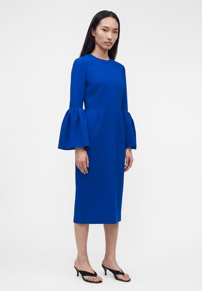 Γυναίκα όρθια με εφαρμοστό φόρεμα μήκους μέχρι τα γόνατα σε βασιλικό μπλε χρώμα με φαρδιά μακριά μανίκια και μαύρα σανδάλια με τακούνι σε λευκό φόντο.