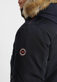 Personne portant une veste d'hiver noire avec capuche bordée de fourrure et un patch rond sur le bras supérieur avec l'inscription « Produits de qualité supérieure ».