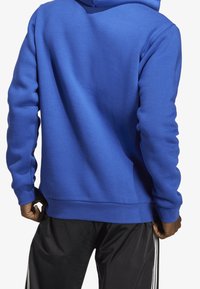 Sininen huppari, joka on valmistettu teksturoidusta kankaasta, rento istuvuus, ribbaatut hihansuut ja yksivärinen muotoilu. Yhdistetty mustiin housuihin.