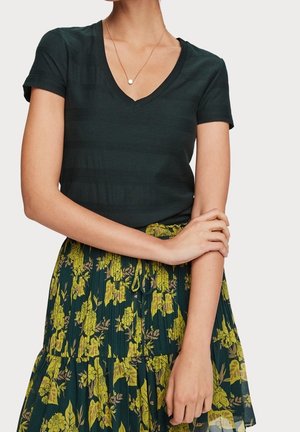 Femme portant un t-shirt vert foncé à manches courtes et col en V, et une jupe plissée avec un motif floral vert et jaune, mains croisées à la taille.