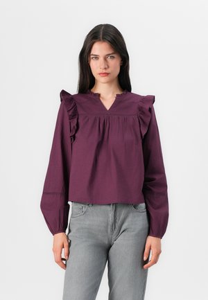 Modellen har på seg en kort, lilla bluse med volanger på skuldrene og V-hals, kombinert med grå jeans; nøytral bakgrunn.