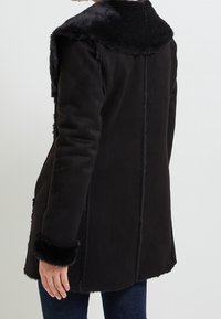 Černý semišový kabát s límcem a manžetami z umělé kožešiny, se rovný střihem a viditelnými švy, což vytváří texturovaný vzhled.