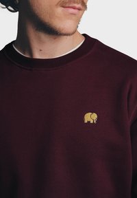 Burgundské triko s kulatým výstřihem a žebrovanými rukávy; má na hrudi vyšitý malý zlatý logo slona. Měkká textilní struktura.