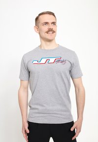 Grå kortærmet T-shirt med et blåt og rødt "JT Racing" logo på forsiden. Stoffet ser glat ud med en afslappet pasform.