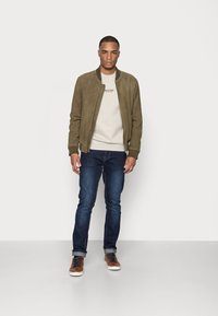 Giacca bomber in suede verde oliva su una felpa beige chiaro, abbinata a jeans blu scuro e sneakers marroni con lacci e suole bianche.