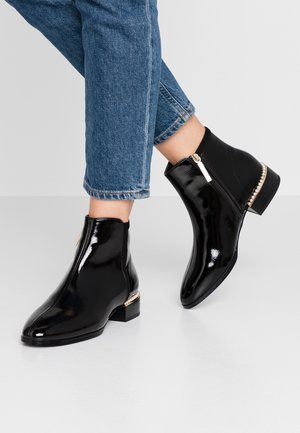Bottines en patent noir avec un bout arrondi, une fermeture éclair sur le côté et un talon bas orné d'un accent doré et perlé.