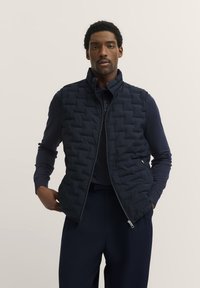 Marineblauer, gepolsterter Steppwesten mit hohem Kragen, gestepptem Muster und Reißverschluss; getragen über einem dunklen Polohemd, kombiniert mit dunklen Hosen.