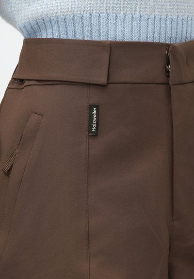 Rudos aukšto liemens kelnes su šonine kišenėmis ir juodu Holzweiler ženklu, dėvimos su šviesiai mėlyna raukšlėta megztine.