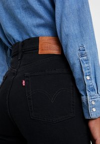 Mustat farkut korkealla vyötäröllä, joissa on ruskea nahkainen Levi's-logopatchi ja punainen tikkipainatus takataskussa. Sininen chambray-paita näkyvissä.