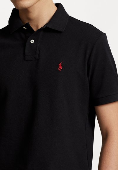 Svart piké skjorte laget av teksturert stoff, med tre-knapps krage, kortermede ermer og en rød brodert logo på venstre bryst.