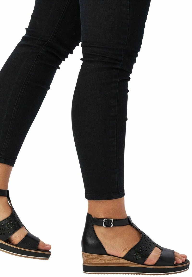 Jambes vêtues d'un jean skinny noir et de sandales compensées noires avec des brides à la cheville et des détails géométriques découpés.