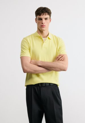 Mladý muž v žltej krátkorukávovej polokošeli a čiernych nohaviciach, stojaci so založenými rukami pred jednoduchým svetlým pozadím.